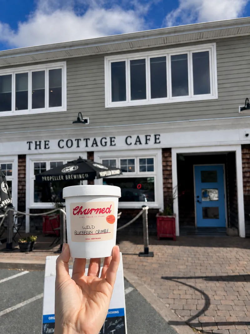 A hand holding a Churned cup in front of the Cottage Cafe storefront and blue door.